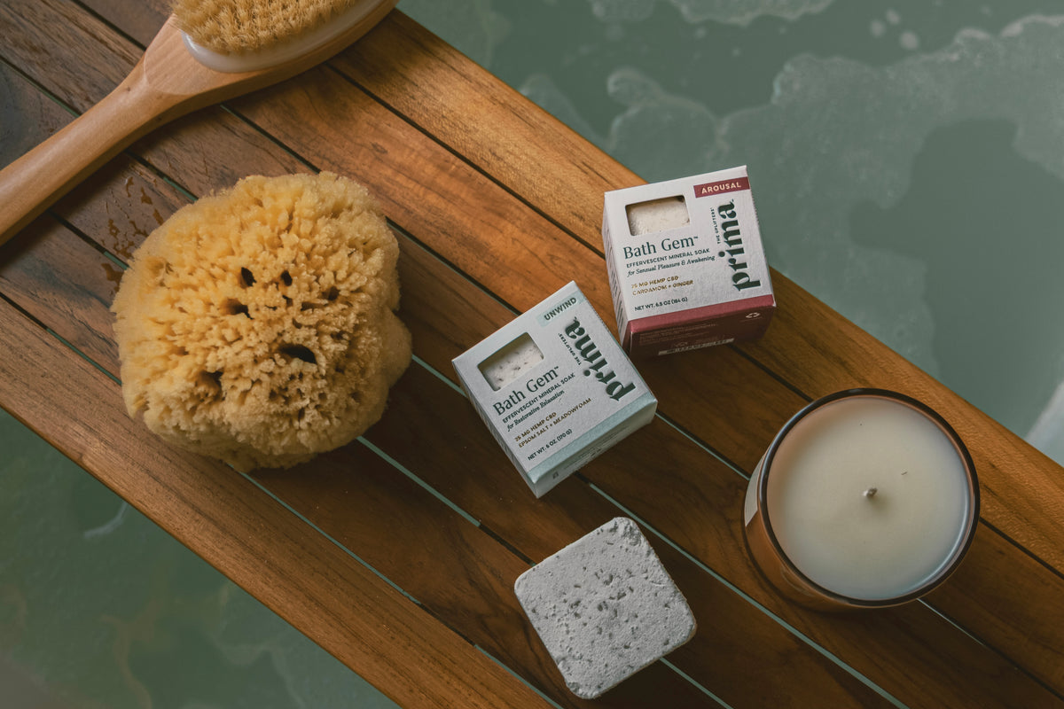A styled flat-lay of bath and body items on a wooden surface: two small Prima Bath Gem boxes, a natural loofah sponge, a gray stone scrubber, and a white candle in a glass jar, with a wooden brush partially visible at the top against a soft green-blue background.